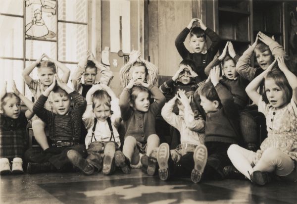 A group of fourteen kindergarten students posed in their classroom, standing and sitting with their arms above their heads. Behind them is a window with a stained glass panel depicting a fairy tale or nursery rhyme character.
