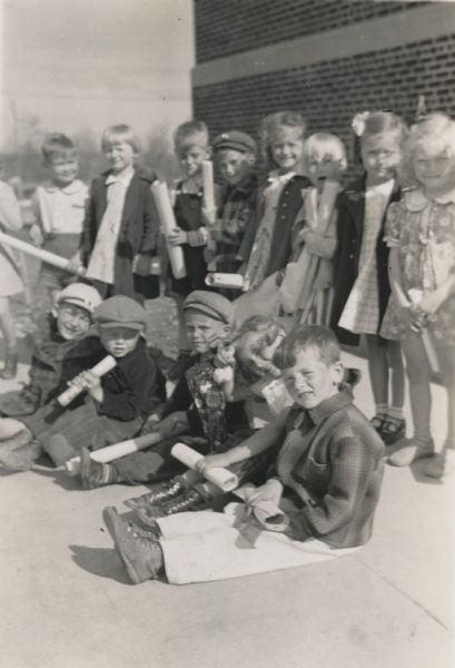 A group of kindergarten students posed sitting and standing outside. Each child is holding a rolled up paper.