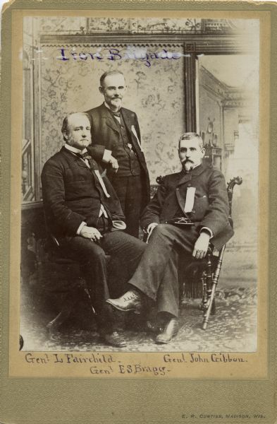 Studio portrait of General Lucius Fairchild, General E.S. Bragg, and General John Gibbon, all of the Iron Brigade. All three men wear an unidentified ribbon on their chests and Gibbon holds a Civil War soldier's cap in his lap.