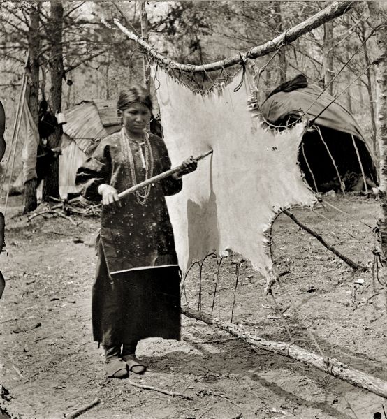 Ho-Chunk woman tanning a buckskin. Typical dwellings (chipotekes) are in the background.