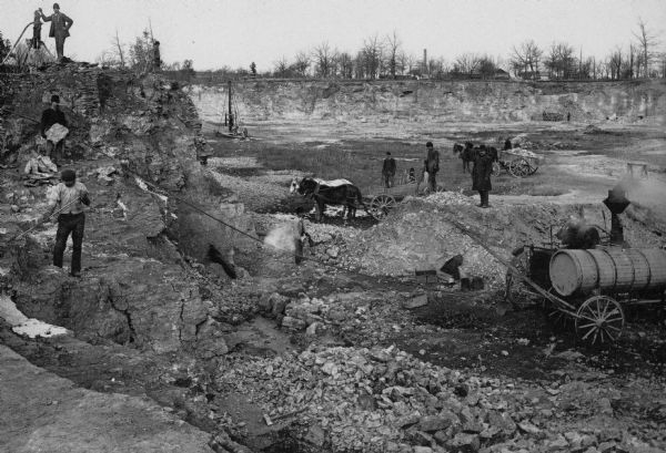Men working in Horlick's stone quarry. Teams of horses pull wagons. There is a steam pump at right. A man in a hat and coat, possibly William Horlick, stands on a pile of stone right of center.