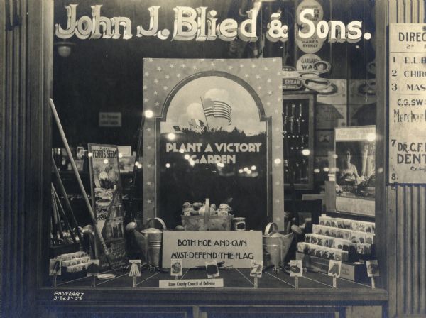 Display in the window of John J. Blied & Sons promoting victory gardens and featuring seed packets, hoes, and watering cans.
