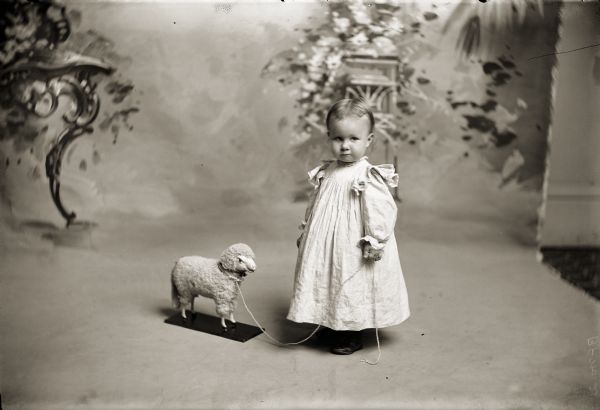 Studio portrait of Ruth Bennett with holding the string of a toy lamb standing in front of a painted backdrop.