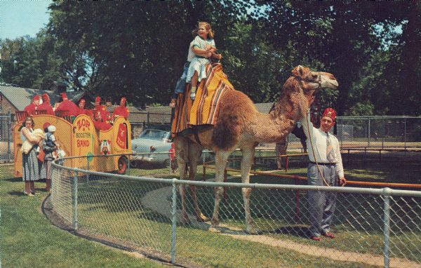 A young girl and another child  riding a camel led by a Zor Shriner at the Vilas Park Zoo (Henry Vilas Zoo). In the background the Zor Booster Band is in a wagon pulled by an automobile. Camel rides were given from 1934 to 2003. Zor Shrine Booster Band wagon is in the background.