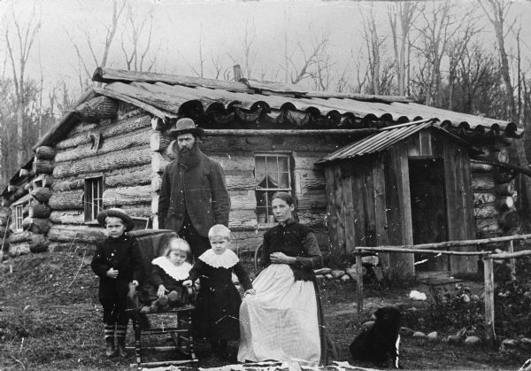 This is a family photograph of Nels Wickstrom with his wife Anna (born Anna Stoel) and their children in front of their log home. The oldest child, wearing the hat, is Nels Odin Wickstrom and the other two are Carrie and Zenella (who later married Reubin Anunson). The child with whom Mrs. Wickstrom is pregnant is Augusta Wickstrom. A dog sits nearby on the right.