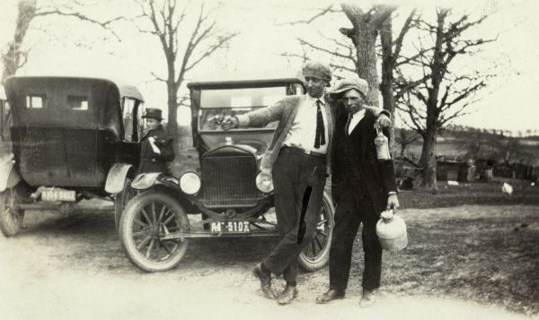 Two men standing in front of two cars, one pointing a handgun at the ground and the other looking at the camera. Each man is holding a bottle, possibly full of illegal alcohol during prohibition. There is a woman in the background between two cars.