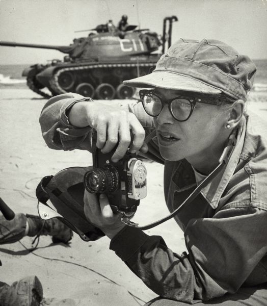 Dickey Chapelle, photographer, on the same Milwaukee beach where she learned to swim as a young girl. She was covering "Operation Inland Seas" celebrating the opening of the St. Lawrence Seaway. She is holding her camera and there is a tank in the background. This is her favorite photograph of herself at work. It was taken by Marine Master Sergeant Louis Lowry.