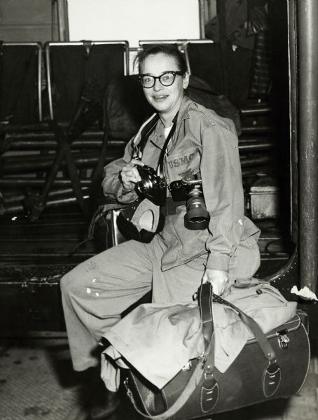 Dickey Chapelle is sitting on the USS "Boxer" with cameras around her neck and luggage at her side.