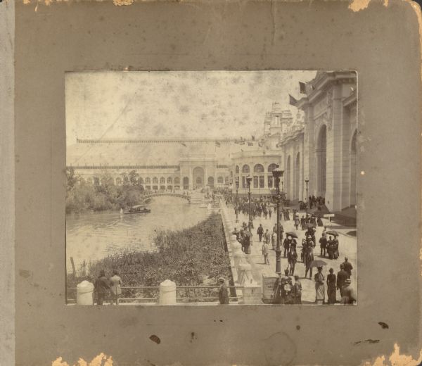 A view of the World's Columbian Exposition mounted on a cardboard album page, looking east from the Transportation Building. On the right are the Mines Building and the Electricity Building, with the Manufactures and Liberal Arts Building in the background. On the left is the lagoon with a motor launch approaching a bridge. There is a drinking water dispenser in the foreground. Women with parasols and men in hats walk the grounds. In the background, a sign advertises a Wagner concert in the Festival Hall.