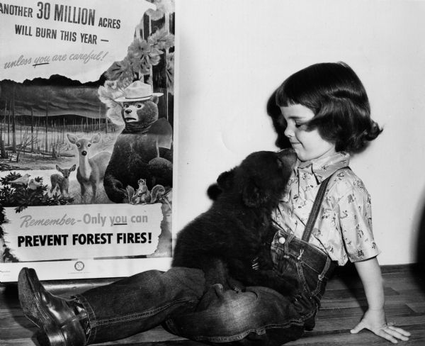 Smokey Bear cub with Judy Bell in front of a fire-prevention poster featuring the character for whom the young bear was named.