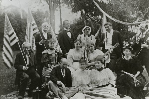 Lucius Fairchild (standing, left), Mary Fairchild (standing, center) and a group of unidentified men and women outdoors in formal attire. Lucius Fairchild and another man each hold a flag, one woman holds a guitar and a woman holds a hand fan.
