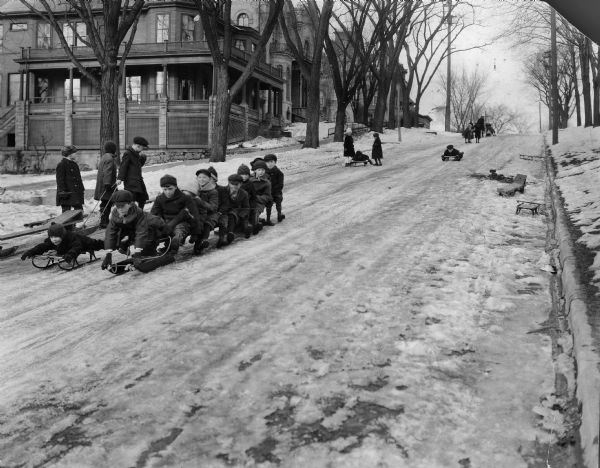 Winter scene with children sledding down 400 block of North Pinckney Street, Madison, Wisconsin, including 10 boys on a coasting bobsled. Three houses are in the background: 414 N. Pinckney the Aylward House built 1900; 424 N. Pinckney the McDonnell/Pierce House (now Mansion Hill Inn) built 1857; and 28 E. Gilman the Keenan House built 1857.
