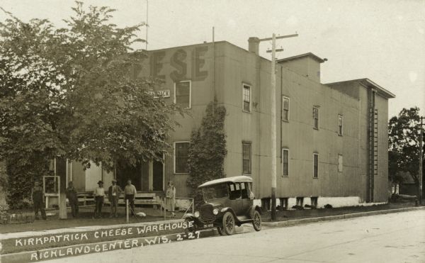 Five men and one woman, possibly employees of the Kirkpatrick Cheese Factory, gathered in front of the factory building. A car is parked along the curb in front of the building. Caption reads: "Kirkpatrick Cheese Warehouse, Richland Center, Wis."