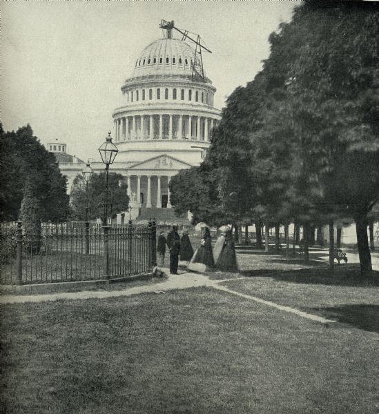 View of the east front of the United States Capitol in Washington, D.C. A small group of people stand near a fence on the lawn in front of the building. A row of trees line a walk at right. There is construction equipment on the dome of the Capitol.