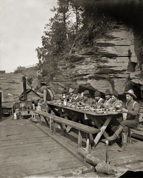 Stereograph of a group of raftsmen eating at a table onboard a lumber raft near the shoreline.