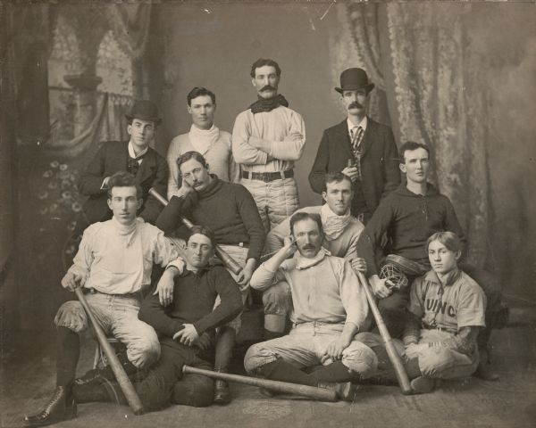 Group portrait of Cooksville Cornhuskers baseball team in front of a painted backdrop. Top left to right: Unknown,  Johnny Sweeney, Tim McCarthy(?), Charles F. McCarthy. Middle section: Unknown, Unknown, Jeremiah McCarthy. Front Row: Dr. Tom McCarthy, D.A. McCarthy, Jim McCarthy and Timothy Hellum.