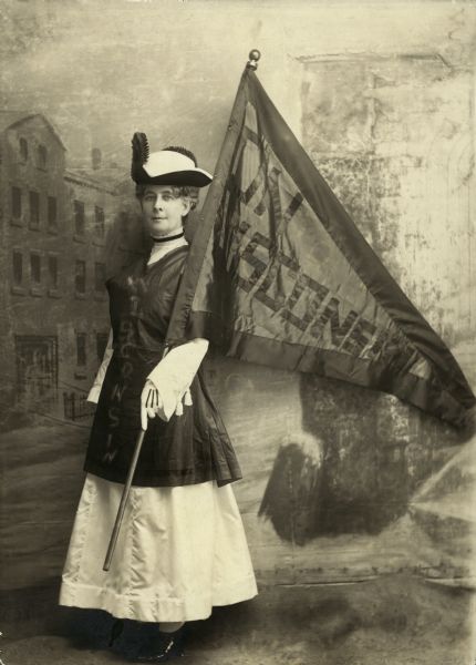 Portrait of Theodora Youmans (Mrs. Henry), President of the Wisconsin Woman Suffrage Association, holding a Wisconsin flag and standing in front of a painted backdrop.
