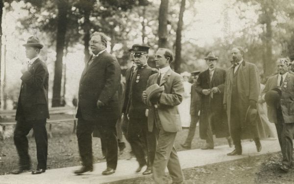 President Taft walking along a path at the Soldier's Home where he addressed veterans. Several others accompany the President, including Mayor David S. Rose, wearing a long coat and holding his hat.