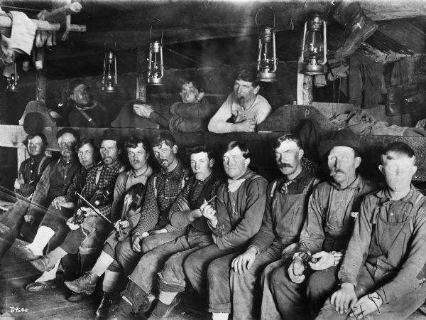 Lumberjacks posing together in a bunkhouse at Ole Emerson's lumber camp. There are lanterns hanging from the ceiling. Two of the men are holding fiddles (violins).