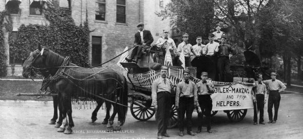Boilermakers and their helpers pose on and around a horse-drawn wagon that bears a sign that reads "Boiler-makers and Helpers," perhaps for a parade. Man of the men are holding hammers. The horses are wearing horse fly bonnets or fly-nets, and the group is posing in front of a large brick building.