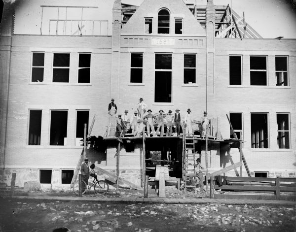 Workers pose on scaffolding in front of the Senior High School during construction. A child and a man with a bicycle are on the lower left side underneath the scaffolding. There is a datestone above the entrance which reads: "1897".