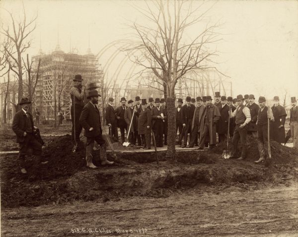 Tree planting ceremony on the Wooded Island during construction of the Columbian Exposition in Jackson Park. Standing to the left of the tree wrapped with twine is George W. Childs holding a shovel. Several other dignitaries and construction workers are gathered around the tree. The framework of a building, possibly the Electrical Building, is going up in the background.