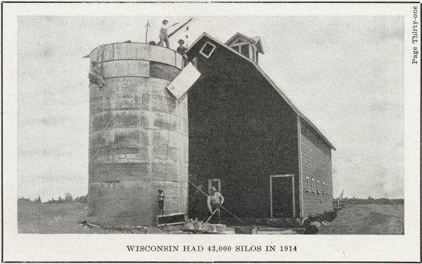 Men using a hoist to lift a panel onto the side of a silo. The silo is next to a barn with a gambrel roof and a cupola.