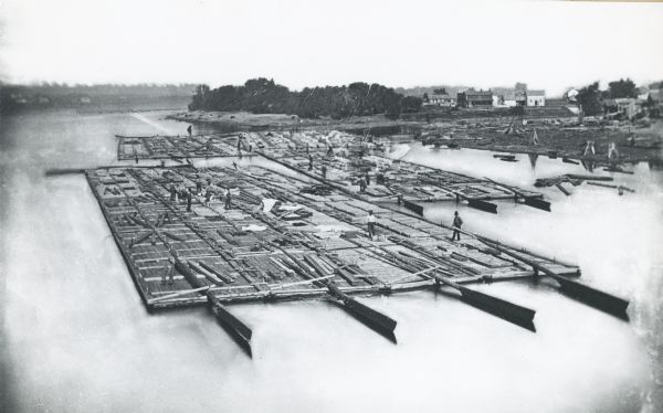 Elevated view of lumber raft and workers on the Chippewa River. In the background on the right are dwellings.