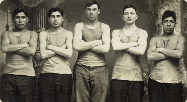 Three-quarter length studio portrait in front of a painted backdrop of five Hayward Indian School students, possibly a basketball team, from L to R: John Coudicon, McBride, Young Joe Gurnoe, unknown, Joe Sharlow. The young men wear tank tops and have their arms crossed across their chests with their muscles flexed.