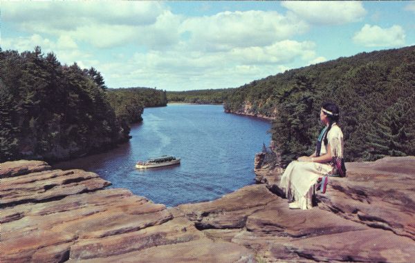 A woman in an Indian dress and headband sits at High Rock in the Upper Dells looking over the Wisconsin River at a tour boat.