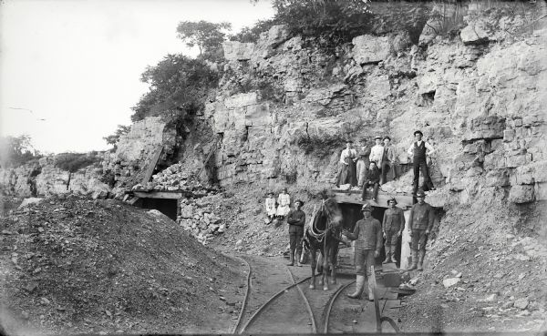 Visitors, dressed in their Sunday best, contrast with the rough garb of the miners. Left to right, miners were: Charles Kanass, Robert "Beaver" Smith (holding the mule), John Smith (his brother), and Charley Klick. The man sitting directly over the mine entrance was Steve Hayes. The man in the dark business vest and white shirt (at extreme right), standing atop the entrance, was James Cundy, mine superintendent. The other men, women and children have not been identified.