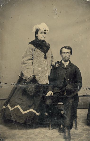 Tintype of Alexander Simplot, sitting, and his wife, standing, who is wearing a fashionable hat and coat and holding a fur muff.