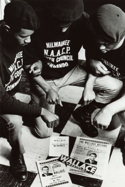 Three members of the National Association for the Advancement of Colored People Youth Council Commandos (Milwaukee) kneel over George Wallace presidential campaign materials. All three young men wear berets and shirts with the name of their organization.