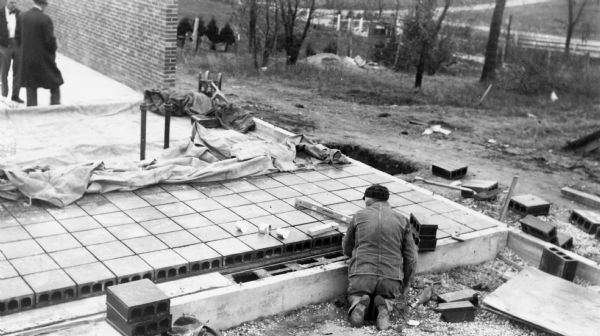 An employee of Clay Products Association builds a floor in a building under construction. Two men stand on the left near a brick wall.