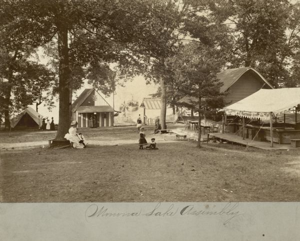 Women and children, who are members of the Monona Lake Assembly, relax beneath the trees at the Monona Lake Assembly grounds. There are tents and small buildings with tables and chairs. In the background is a shoreline with a boat on the lake near a pier.