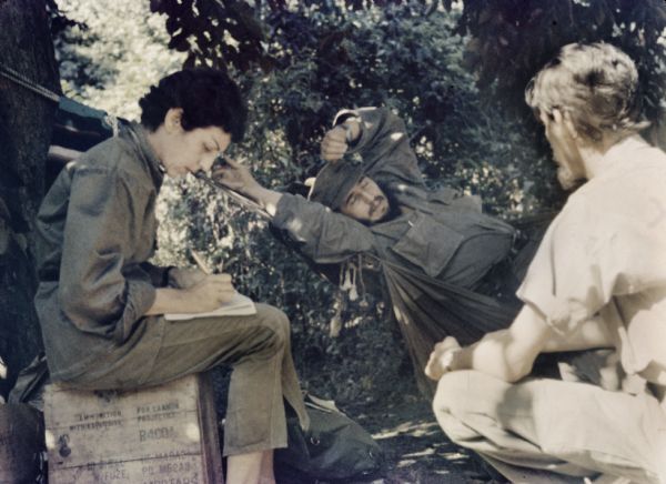 Informal meeting in the woods in Oriente Province during the Cuban Revolution. Fidel Castro is reclining on a hammock talking to a crouching man in the foreground. Celia Sánchez is sitting on a wooden crate writing.