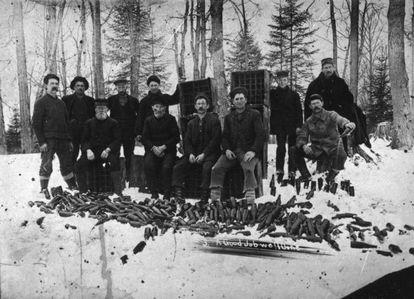Group of men posing in a wooded area among liquor bottles that have been emptied into the snow, the result of a raid on a saloon in the town of Polar which was operated in defiance of the town's no-license administration. A man at left pours the contents of a bottle into the snow.