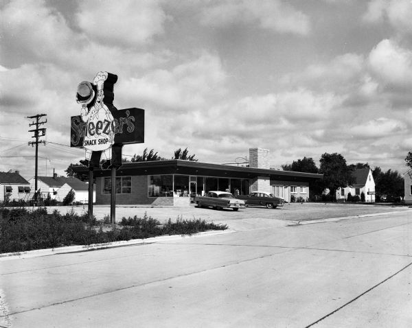 Exterior view from road of Sneezer's Snack Shop with two cars in the parking lot. The neon sign features a chef holding a hamburger on a plate.