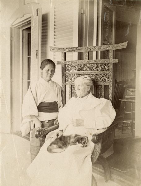 Eliza Catherine Scidmore seated in an ornate chair with a small dog curled up on her lap. Next to her stands a Japanese woman.