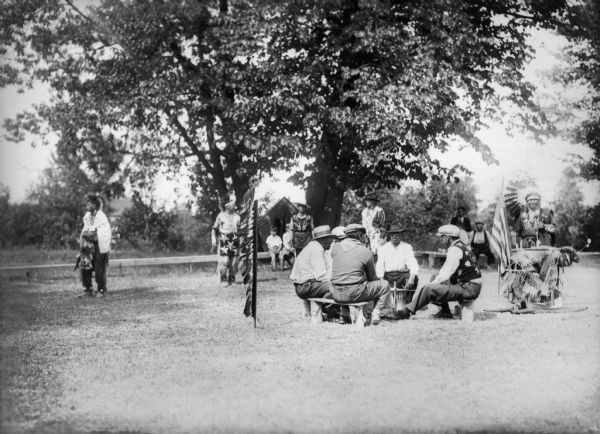 View of five men wearing hats sitting outside in a circle around a drum while six other men dressed in Native American clothing are standing behind the seated men. Two boys on the left and two men on the right are sitting on benches in the background, with trees and shrubs visible behind them. Off to the right is an American flag and a Native American peace pipe.