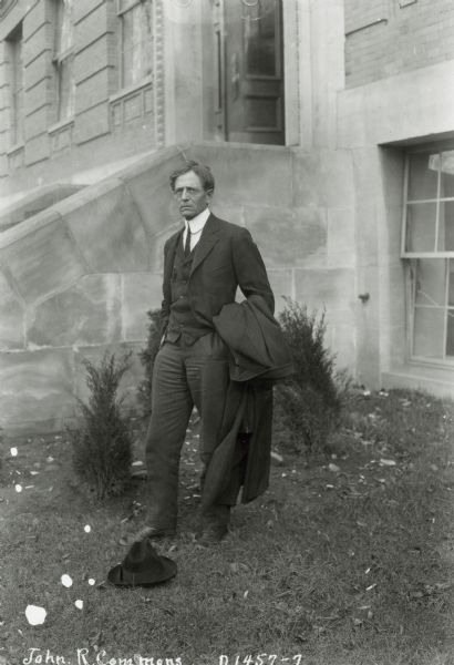 John R. Commons, a University of Wisconsin professor, is shown standing outside Sterling Hall with his hat on the ground at his feet.