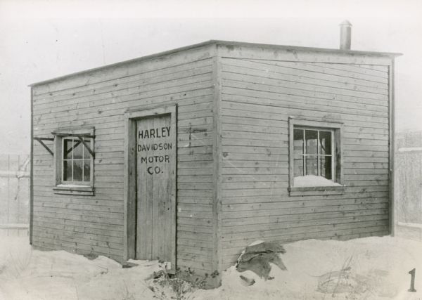 The original shop in the Davidson family backyard where the first Harley-Davidson motorcycles were assembled. There is snow on the ground. The sign on the door reads: "Harley Davidson Motor Co."