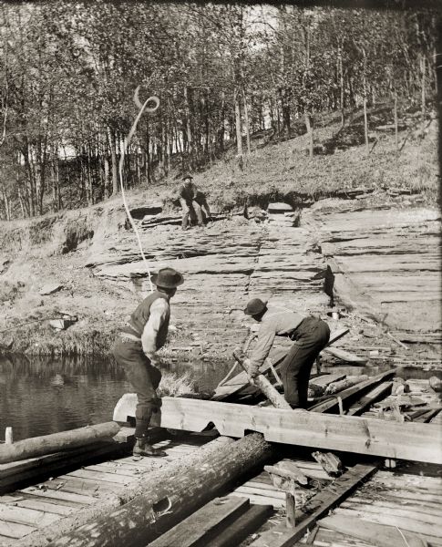 Two men are on a raft. One man is bending to lift a piece of wood, and the second man is throwing a rope to a third man on the shore.