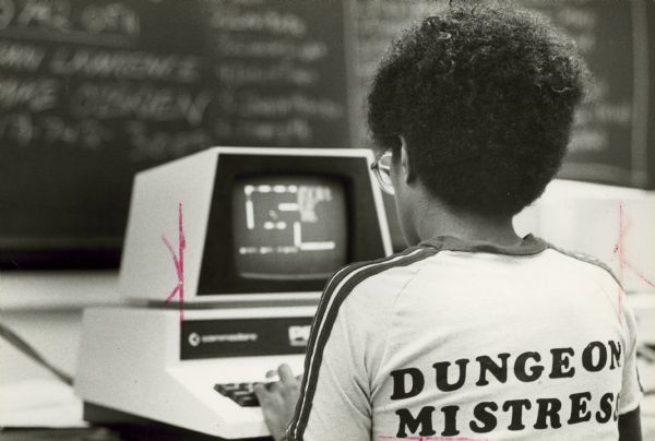 View from behind of a young woman wearing a t-shirt with the title Dungeon Mistress printed on the back while she plays an adventure game on a computer. In the background is a blackboard