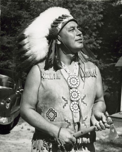Image is an outdoor portrait of a performer wearing a fringed vest, beaded necklace and headdress, and holding a small axe. Member of the Peavey Falls group of dancers and musicians from the Menominee Indian Reservation in Wisconsin.