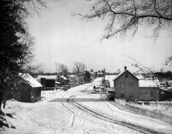 Elevated winter scene of snow-covered road through Keshena, Wisconsin. There are horse-drawn vehicles and pedestrians, and a church is in the background.