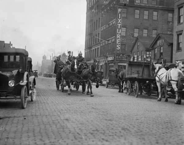 Horse-drawn fire wagon coming down a cobblestone street in Milwaukee. The fact that automobiles are also in the photograph suggests that the transition to motor-driven emergency vehicles did not immediately follow the introduction of passenger cars.