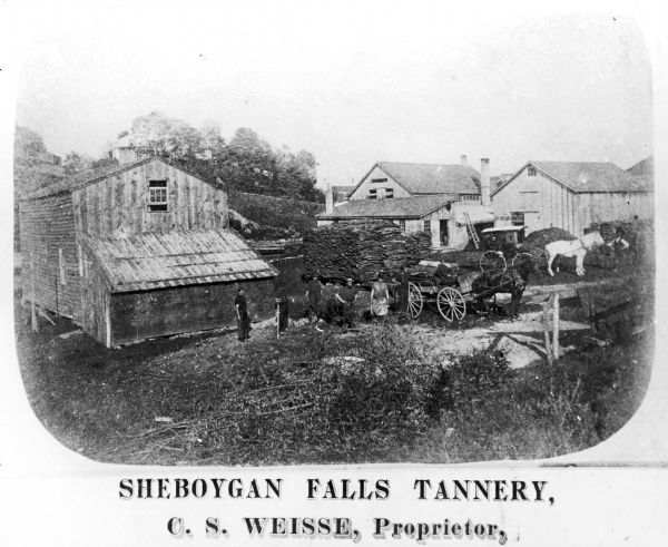Elevated view of several buildings making up the Sheboygan Falls Tannery, which was owned by proprietor C.S. Weisse. Weisse is at the far right, standing with a white horse. Herman Sadler is standing by the wagon wheel. Mr. Kobul is wearing an apron and Mr. Busse is second from the left.