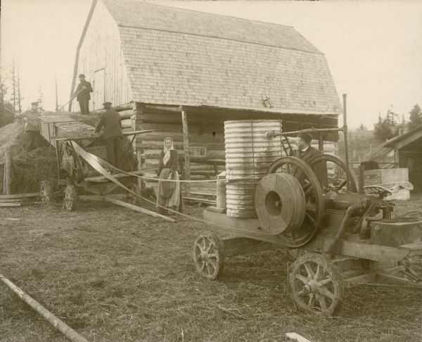 Men operating a hay press powered by an International Harvester stationary engine as a woman is looking on. They are working near a barn which has chink-log construction.