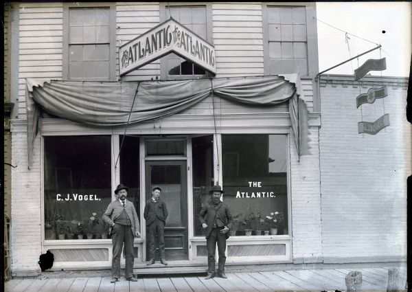 Two older men and one younger man standing on a boardwalk in front of the Atlantic Saloon owned by C.J. Vogel. A hanging sign to the right advertises for Schreihart's Brewing Co. and also bears the name of C.J. Vogel. The center sign is undecipherable.